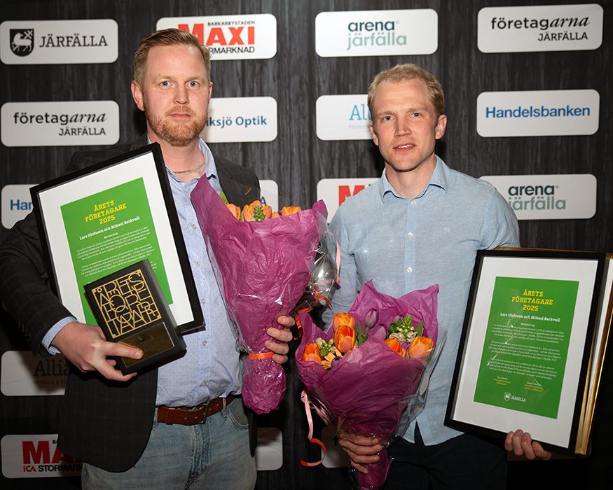 Två män med blombuketter och inramade diplom framför en vägg med sponsorlogotyper