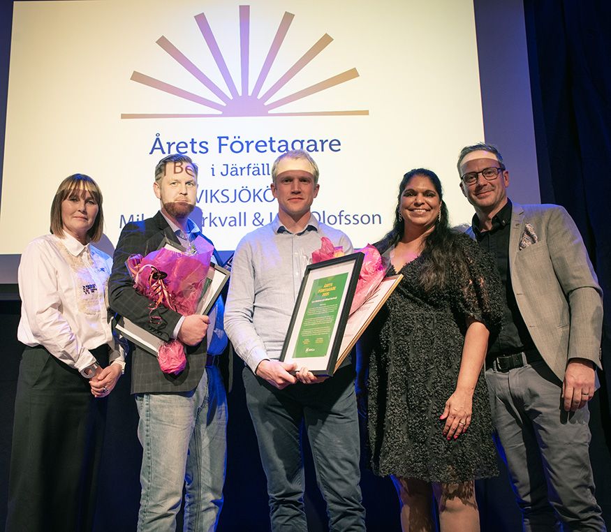 Fem personer på en scen framför en skärm med texten Årets Företagare i Järfälla. Två av dem håller diplom och blommor.